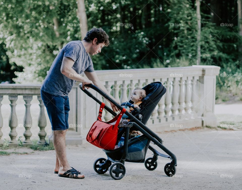 Handsome young male father in a t-shirt and denim shorts with curly brown hair reaches out his hand to his little daughter sitting in a stroller and looking tenderly at him in a public park with a blurred background, close-up side view. Fathers conce