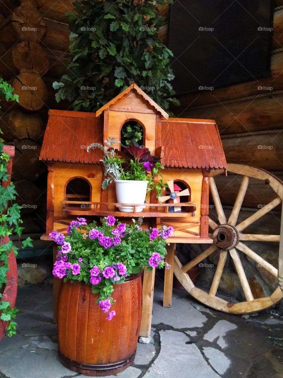 A wooden house, a wooden wheel, a wooden tub in which purple petunias grow. purple and wood contrast