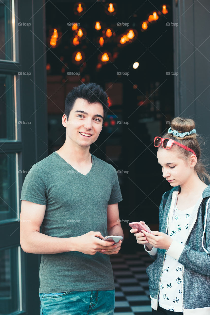 Couple of friends, teenage girl and boy, using smartphones, talking together, standing on street in center of town, spending time together
