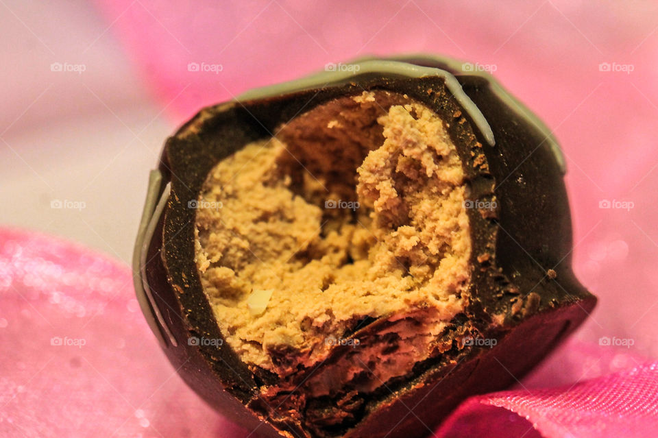 Macro of a half-eaten chocolate sitting on a sheer, glittery pink ribbon. The chocolate is a truffle which is a soft ganache formed into a ball & enrobed in a dark chocolate shell of hardened chocolate with a drizzle of hardened white chocolate. 