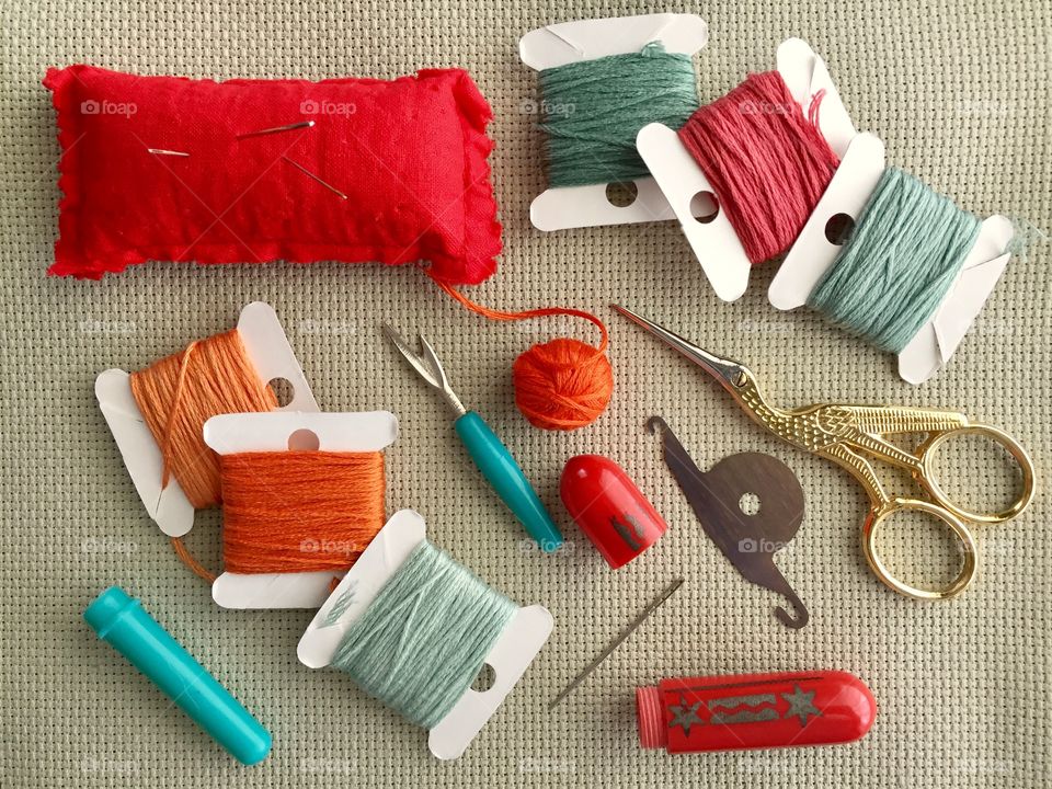 Arts and Crafts Supply - flat lay of cross stitch fabric, pin cushion with needles, needle case, embroidery floss on cards, ball of embroidery floss, decorative gold bird scissors, threader, ripper