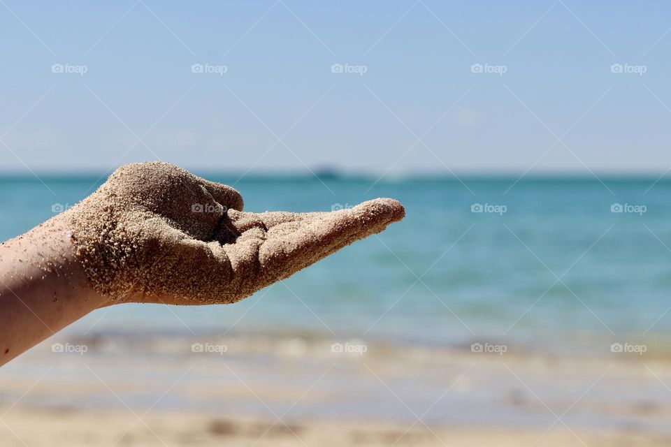 Human hand with beach sand outstretched against backdrop of turquoise ocean horizon at beach on sunny day in south Australia, copy text space