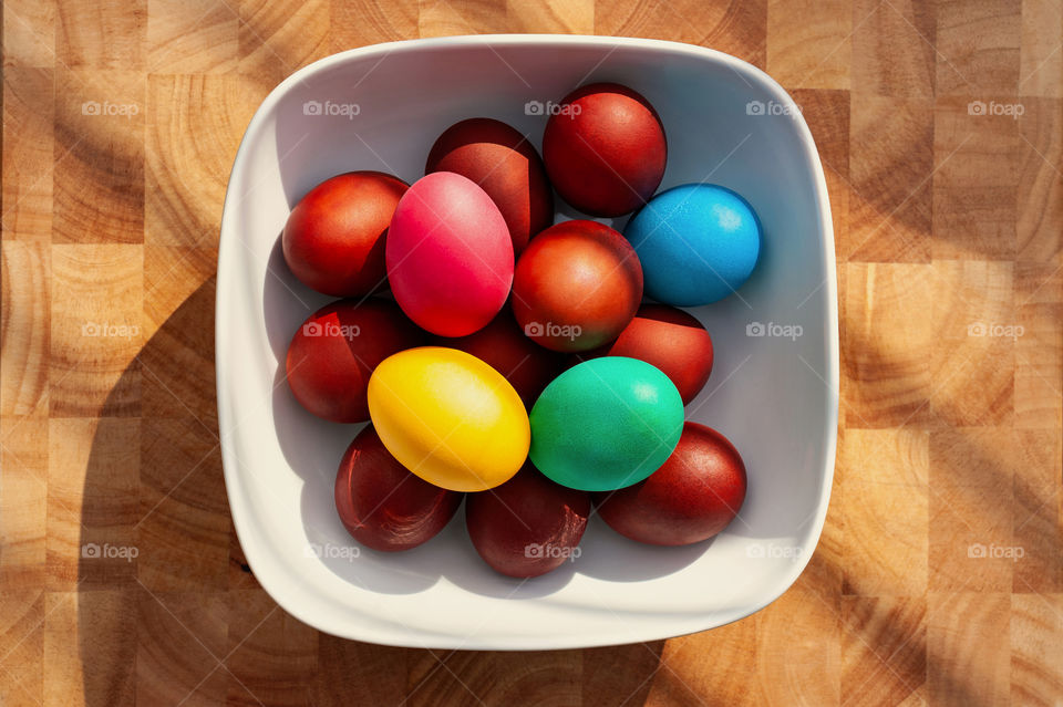 Colorful Easter eggs in white square bowl on wooden background.