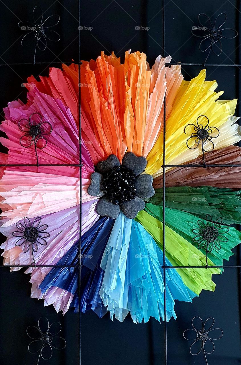 Flatlay composition of a multi-coloured flower made of silk papers, heart-shaped pebbles, black beads and black metal flowers placed on black paper, creating a colour explosion