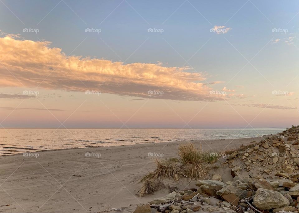 Liquid Fresh water Lake Eerie on Cedar Point shoreline and sandy, rocky beach with purple, pink and blue sunset sky divided by clouds. Beach grass and rocks in foreground