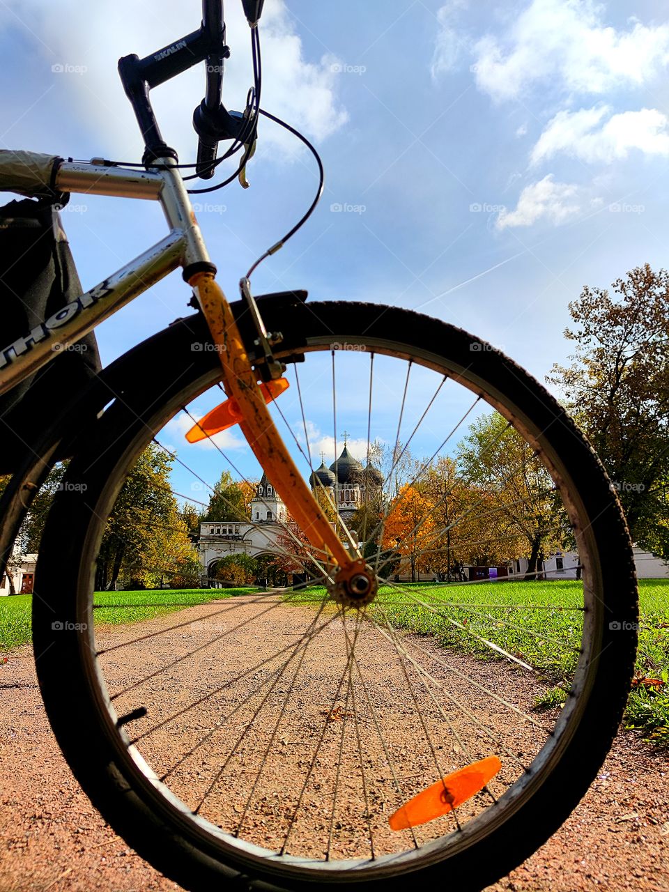 Green lawn with benches.  Multicolored autumn trees.  Ancient fortress wall with a white tower. Domes of an ancient church. Blue sky.  Bicycle wheel in the foreground