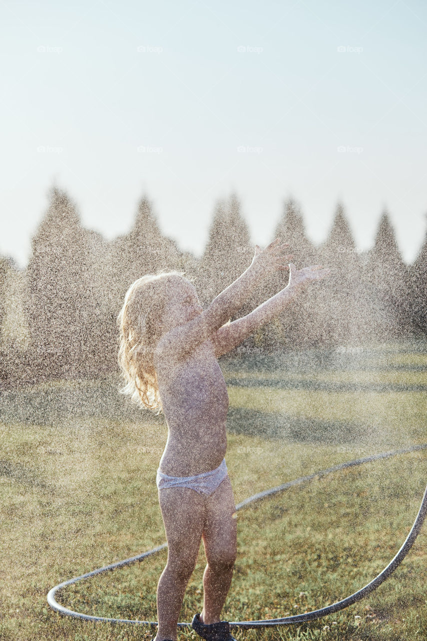 Little cute adorable girl enjoying a cool water sprayed by her mother during hot summer day in backyard. Candid people, real moments, authentic situations