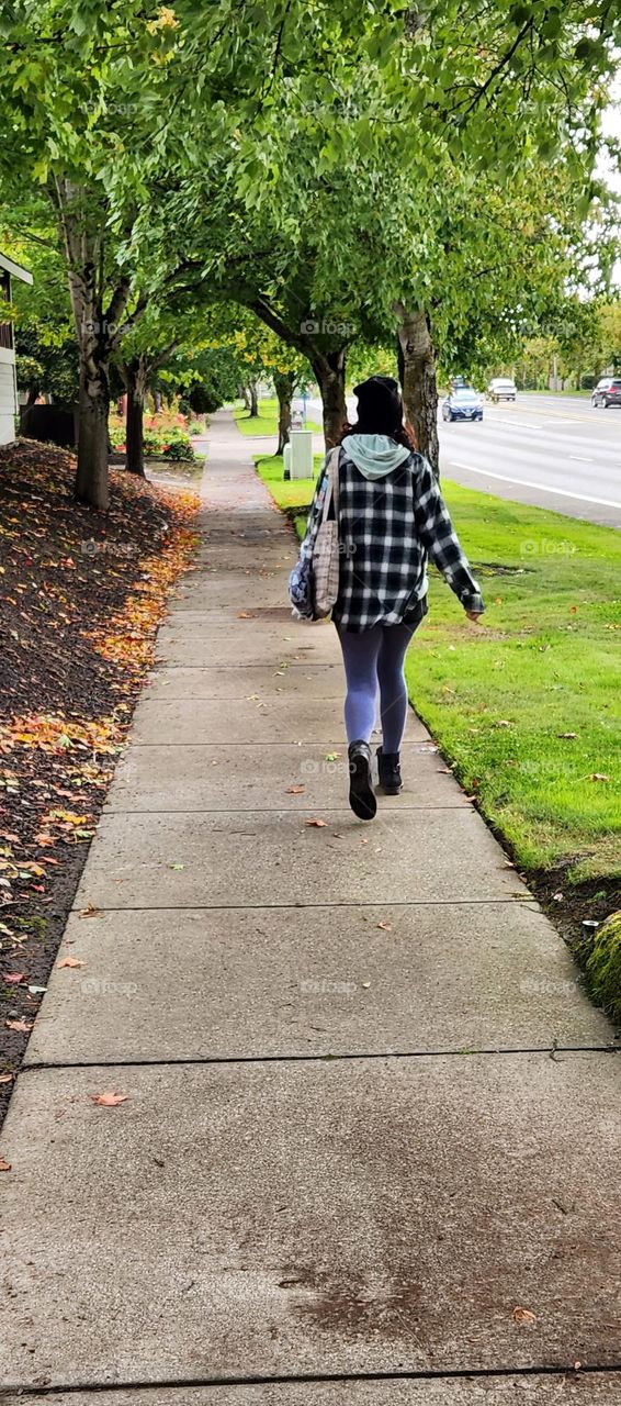 A young woman dressed in warm clothing walking down an Oregon suburban sidewalk lined with fallen Autumn leaves