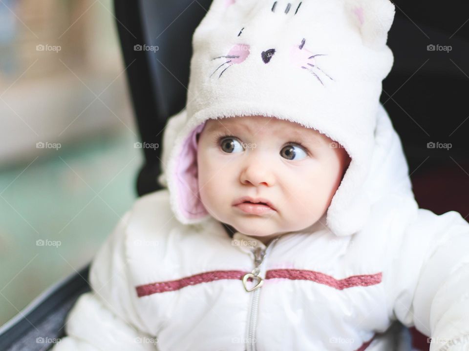 Portrait of a beautiful little Caucasian baby girl in a winter hat, jacket with a very surprised and curious emotion on her face sits in a stroller and looks to the side, close-up side view. Surprised emotion concept,