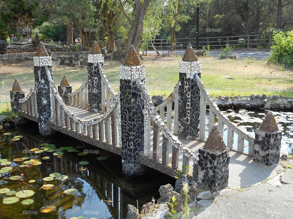 A beautiful and ornate rock bridge on a peaceful path crosses a fairytale style mote with lots of Lillie Pads at Peterson’s Rock Garden on a sunny summer day in Central Oregon.