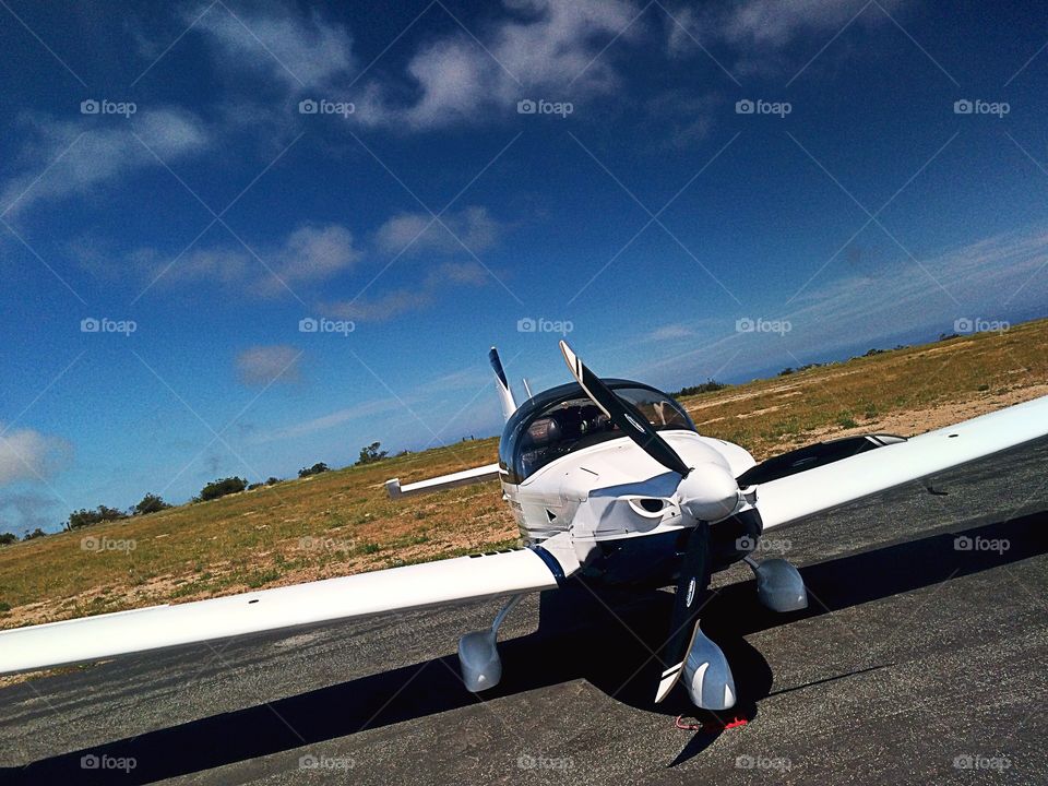 The front view of a small, low-wing private airplane on the tarmac at Catalina Island with clouds and blue sky in the background. Plane is slightly tilted. 