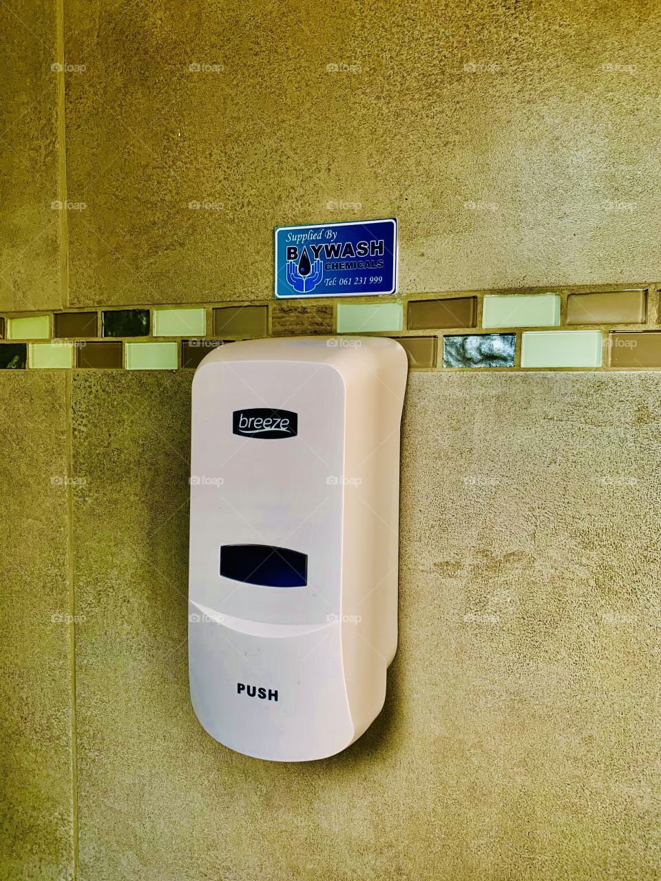 A white bathroom soap holder on the tiles covered wall in a restroom for hand washing. With a blue paper sing indicative of hand hygiene.