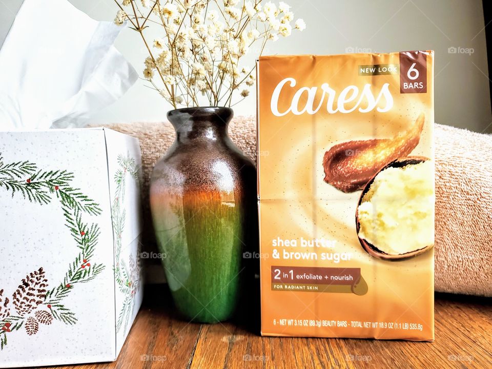 The new look Caress shea butter and brown sugar six pack of bar soap on a wood surface next to a matching vase with florals, a tissue box and tan towel.