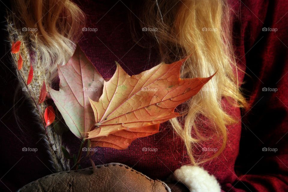A woman in a dark red shirt and red-blonde long hair holds autumn leaves and grass in her hand in brown fur gloves