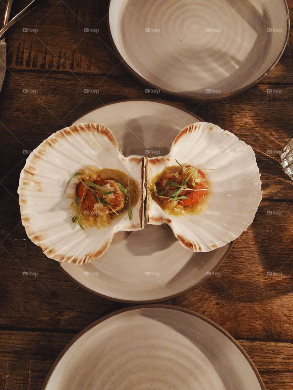 Two fresh scallops served in their shells on earthenware plate and wooden table in restaurant in London, England.