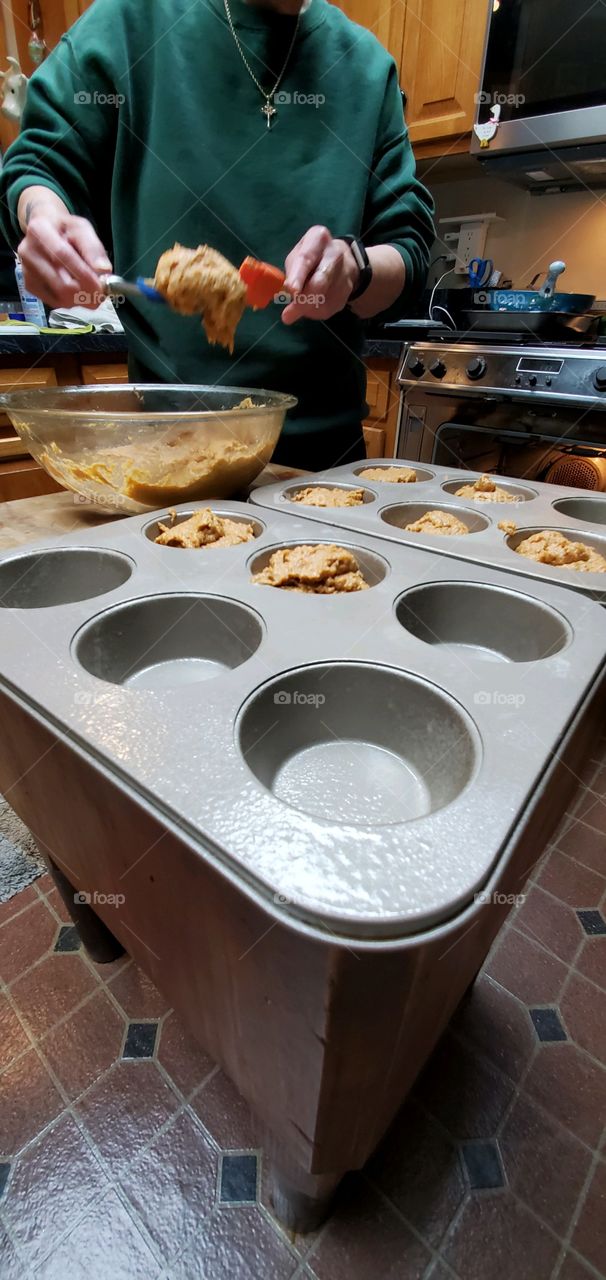 #stayathome-cooking. Hands using utensils to fill muffin pans with homemade batter and then bake in open oven that's waiting.