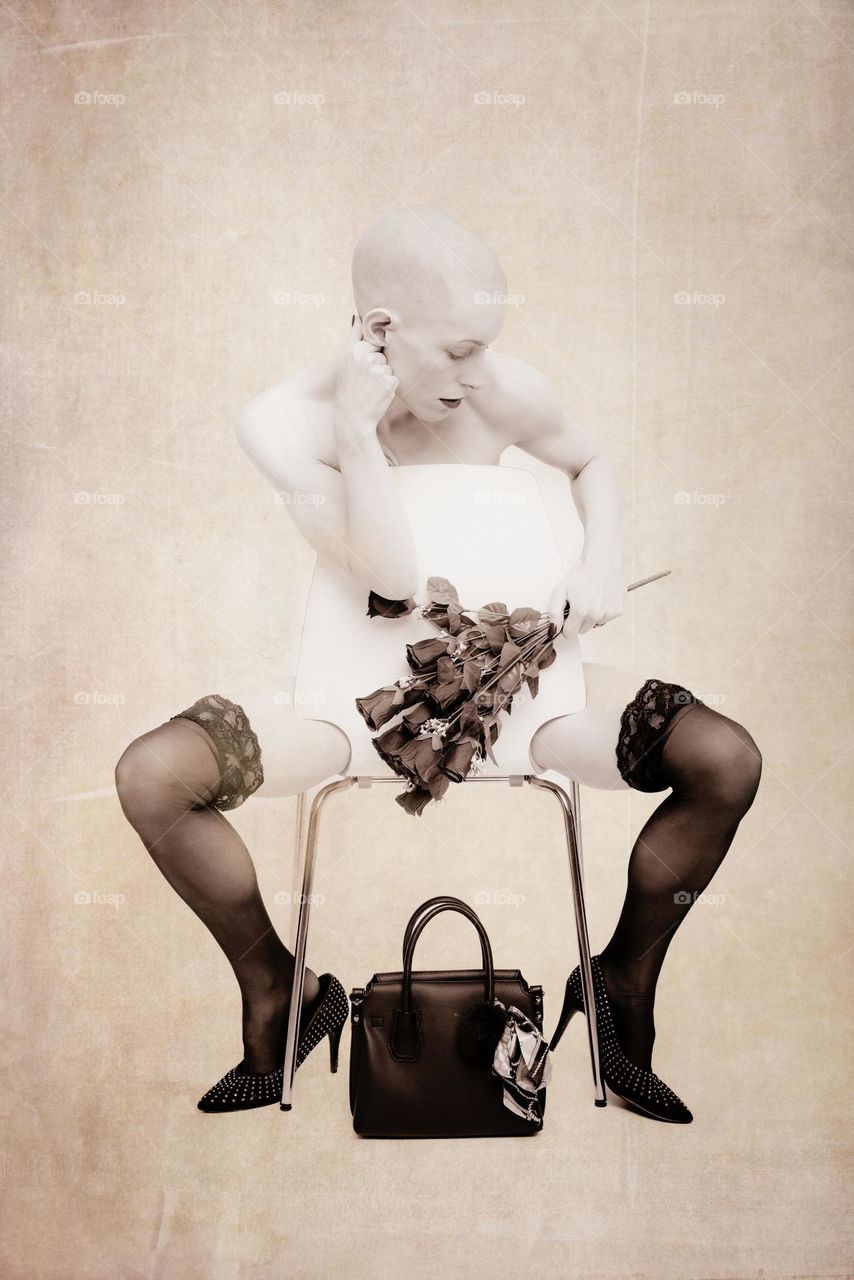 Sepia toned image of girl seated wearing only stockings and high heeled shoes. Her top half is obscured by the chair back. She is holding black roses and her handbag is on the floor