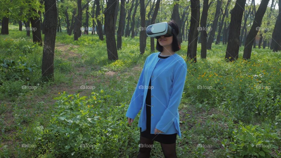 Portrait of trendy woman in blue coat using virtual reality glasses simulator in the park or forest at spring 