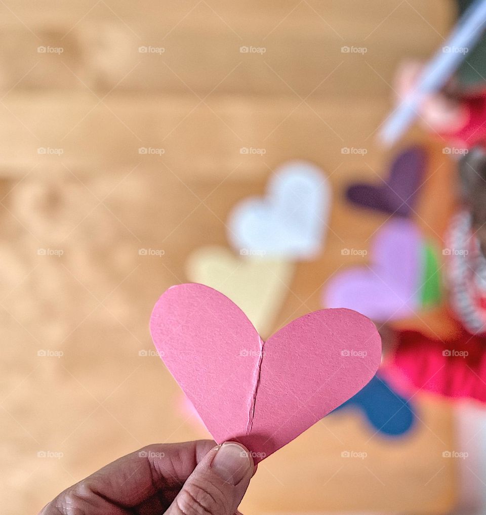 Holding a pink heart while child colors on others, making valentines for family, mailing out valentines to friends and family, spreading love with crafts, pens and paper, crafting indoors, winter time crafts 