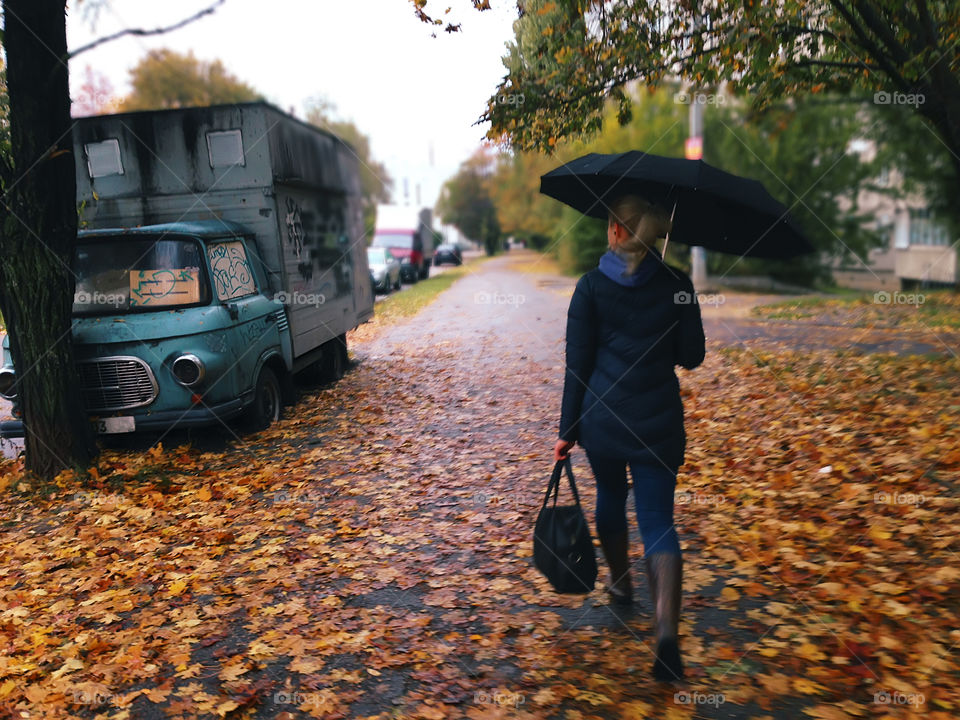 Young woman with headphones walking by colorful fallen autumn leaves at the city park road under umbrella 