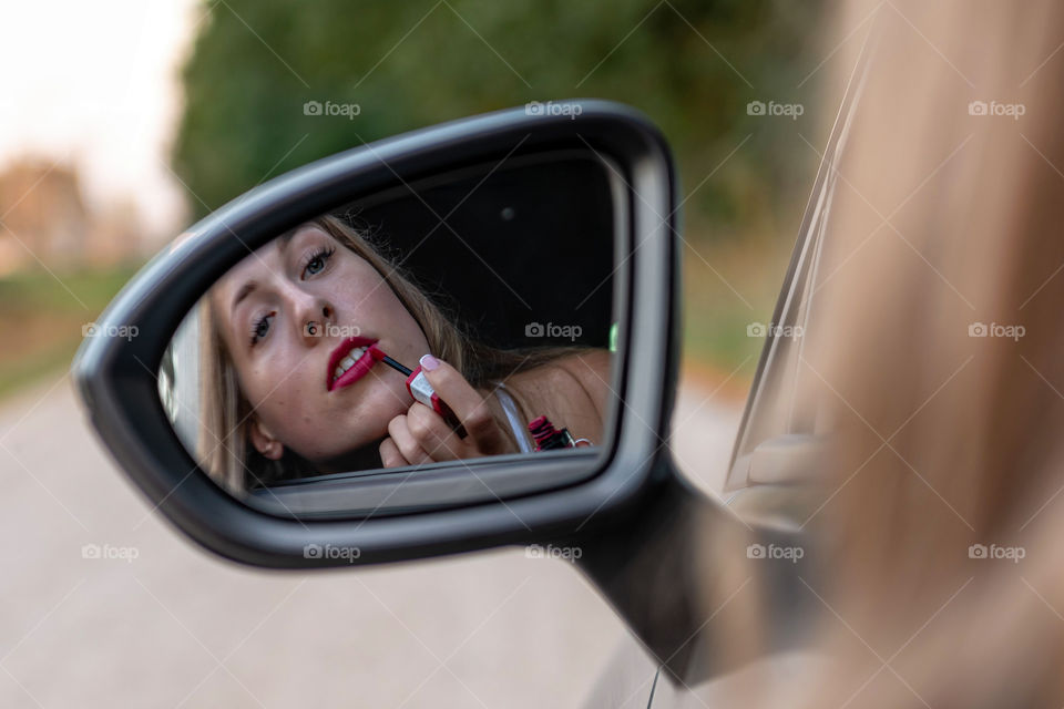 A young, beautiful woman with long hair looks in the car rear view's mirror and paints her lips.