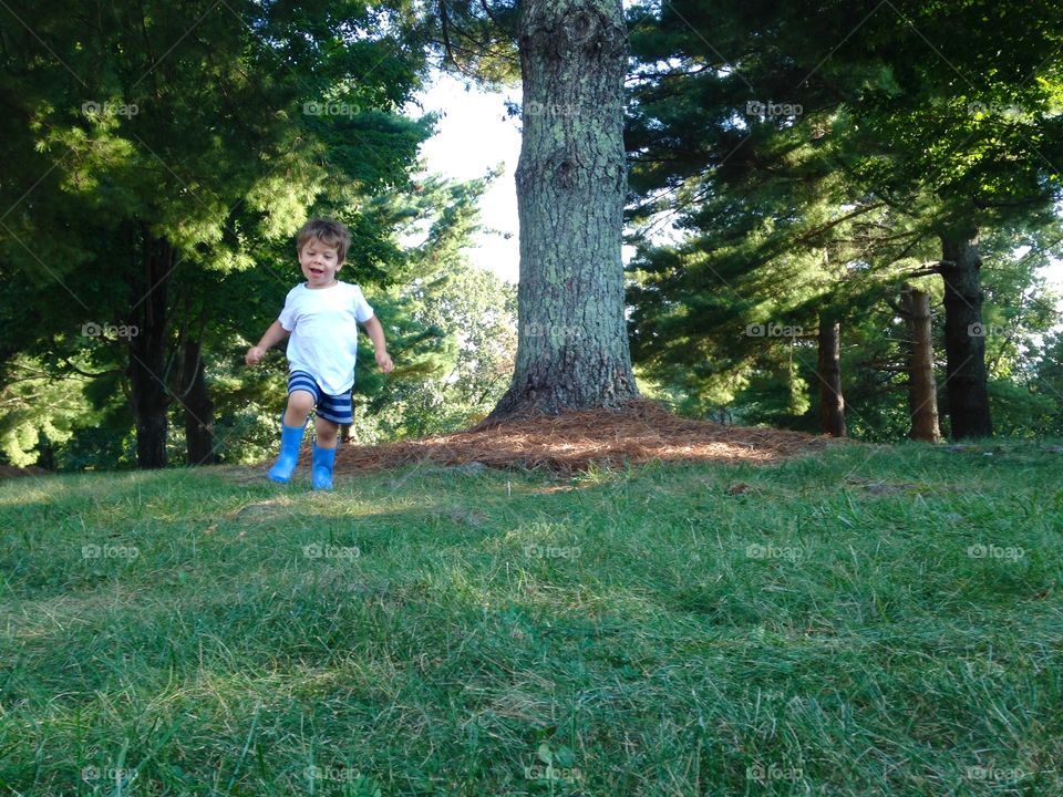 A young boy running and playing outside in the grass in the summer with rain boots on.