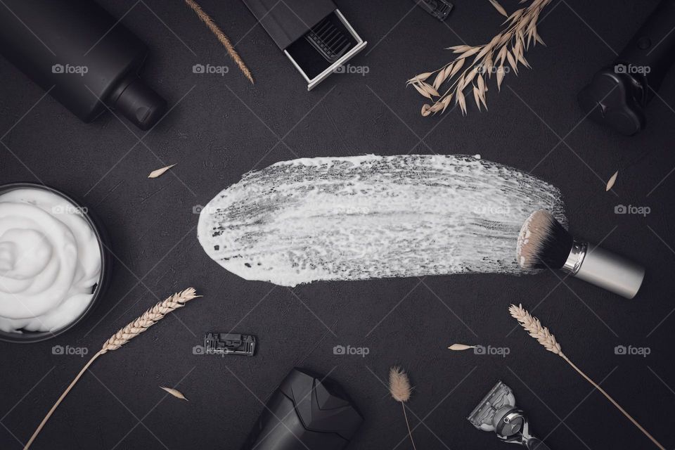 A set of cosmetic products for shaving the beard: razors, brush, shaving foam, electric shaving machine, aftershave, combs and dried flowers with a dab of foam on a black background, flat lay. The concept of a barbershop man, shaving man, beard care.