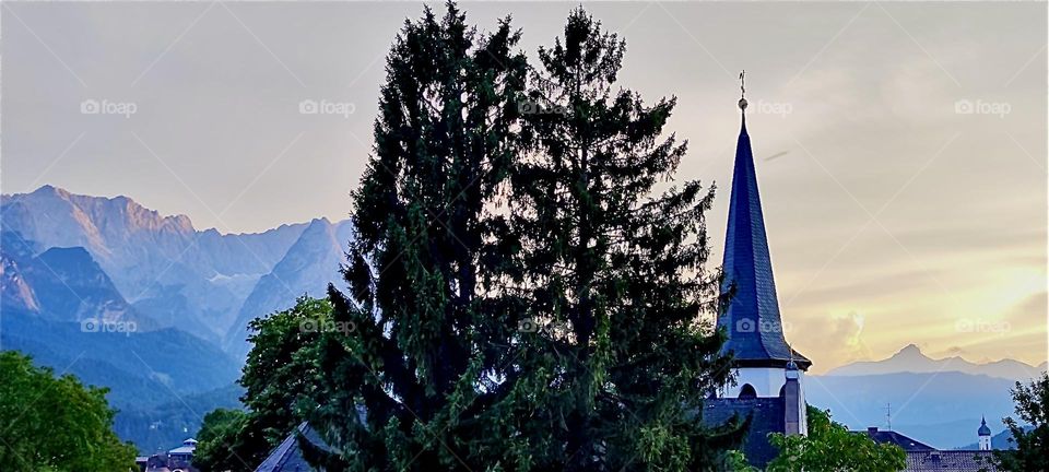 A beautiful silhouette of tall evergreen trees against a church steeple emerges at “Garmisch-Partenkirchen” in the Bavarian “Alps” close to the “Zugspitze”, Germany’s highest mountain at 2962 m. 2024. Hypnotic Productions