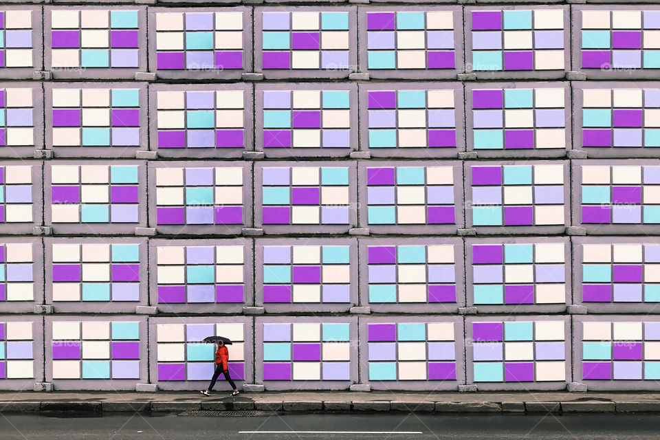 Tiny human walking with umbrella by the colorful wall 