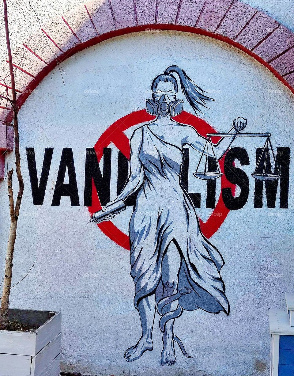 Urban art.  Graffiti.  A blindfolded woman holds a scale.  A snake is crawling on the woman's leg.  The word "VANDALISM" and a red stop sign are written in the background