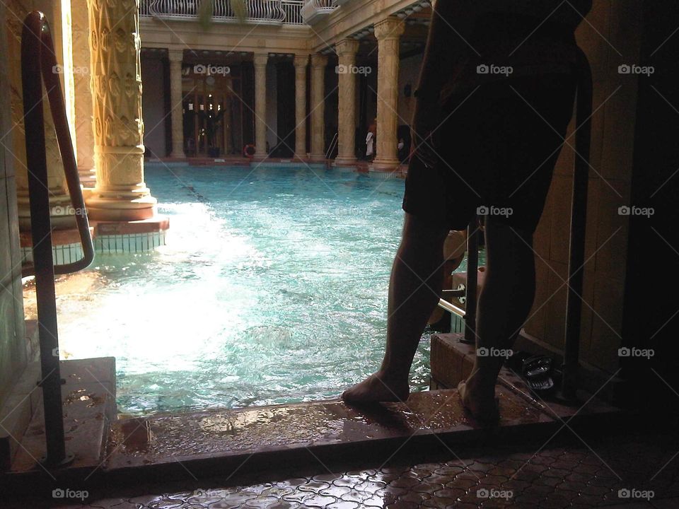 male about to enter an indoor swimming pool with sunlight illuminating the water's surface.