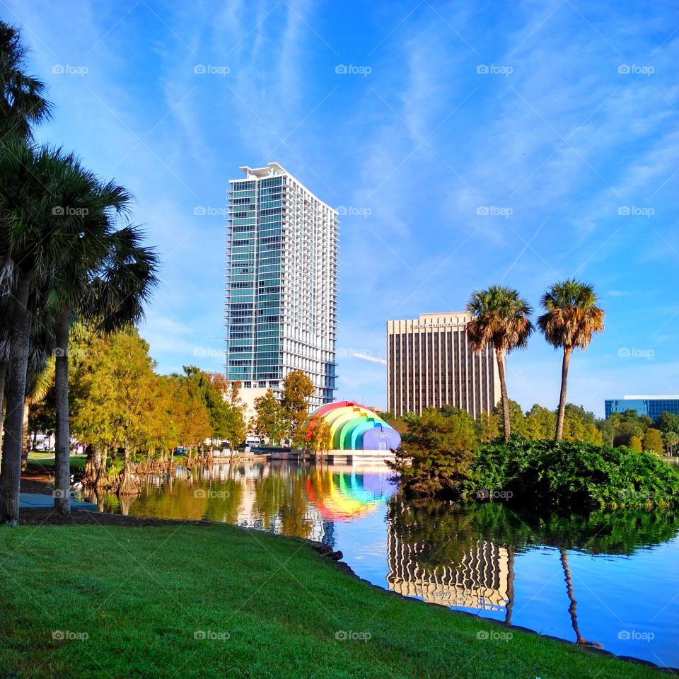 A landscape photo of Lake Eola in Orlando, Florida. Shown are the lake, the rainbow amphitheatre, trees ,and part of the city.