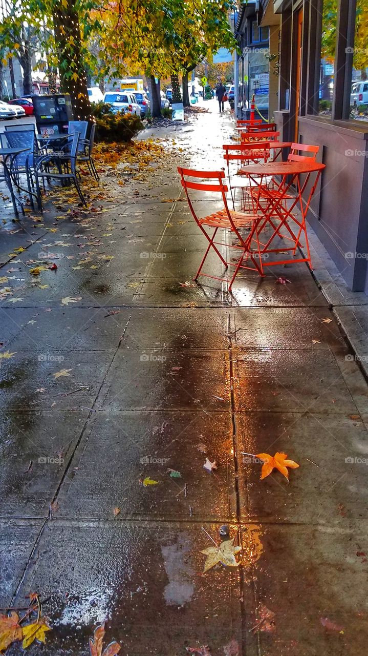 You can see the reflections of the trees on the sidewalk after a rain next to the tables and chairs in front of a restaurant.