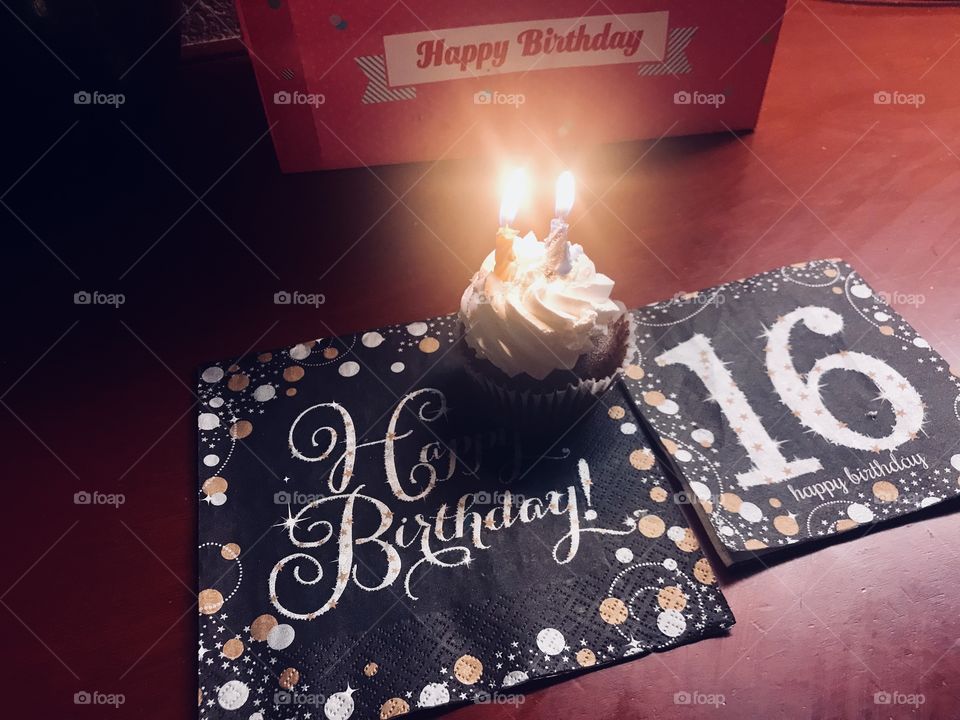 A display of happy birthday napkins for a sweet 16 party with a cupcake made of chocolate and vanilla frosting with candles on top glowing brightly next to a hot pink gift bag.