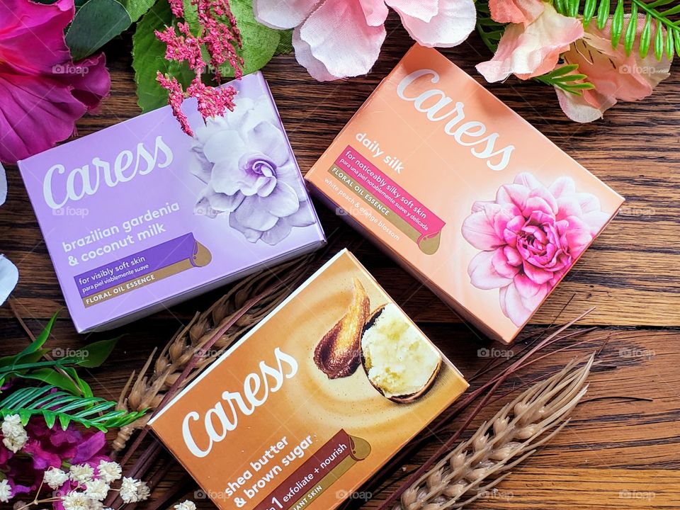 The new look Caress bar single packs: Daily silk, shea butter and brown sugar, and Brazilian gardenia & coconut milk on a wood surface surrounded by matching florals.