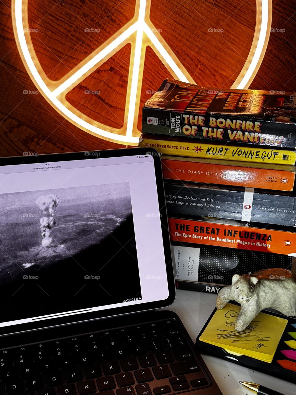 Desktop with iPad showing Hiroshima bomb take from aircraft window, near stack of books, with trendy peace sign tube light in background and handmade polar bear figure standing on Post It Notes pad with pen