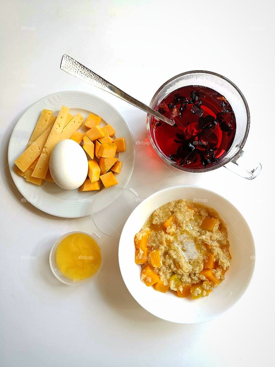 Breakfast.  Plate with oatmeal porridge and pumpkin pieces.  A jar of honey.  Plate with chicken egg and cheese slices.  Glass of cardamom tea
