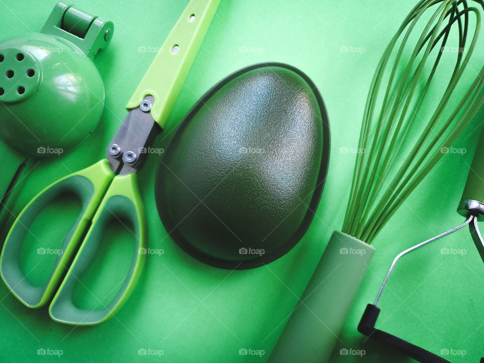 Five green kitchen utensils placed in a diagnol fashion: lemon/lime squeezer/press, herb mincing scissors, avocado holder container, silicone coated wire wisk, and a masher.