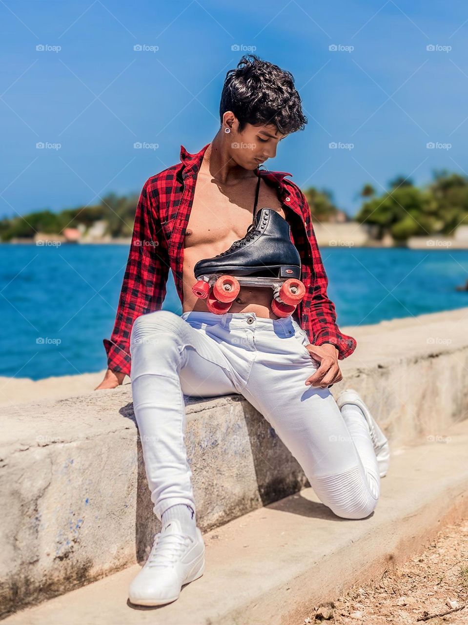 Portrait of a young man sitting on the beach against a background of sea and blue sky. The person was wearing casual clothes and holding black roller skates with red wheels