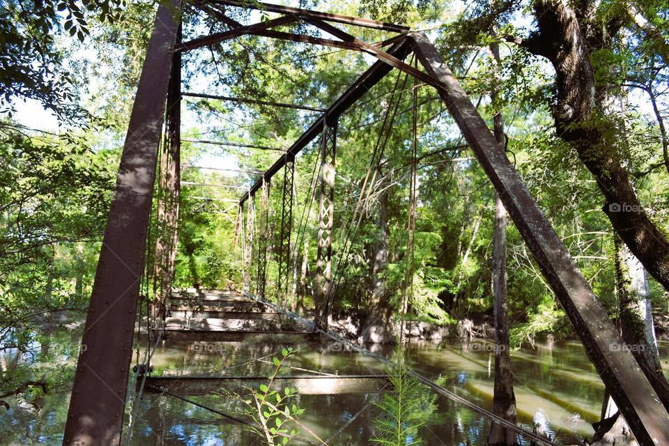 An old metal bridge with no bottom surrounded by lush green trees over murky water in the daytime