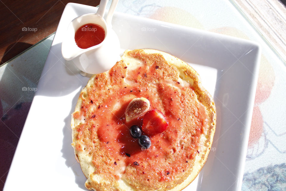 Pancakes with fruit a strawberry sauce, shot from above on a square white plate, with a jug of sauce 