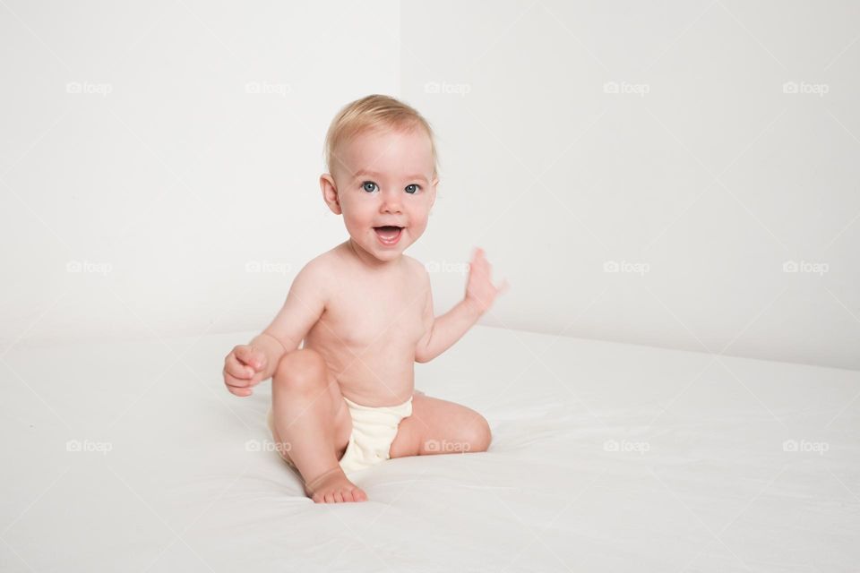 Happy smiling baby sitting on white bed in white bedroom