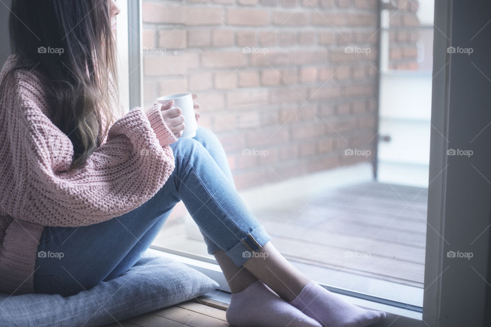 A woman wearing a cozy sweater and holding a cup and sitting near the window 