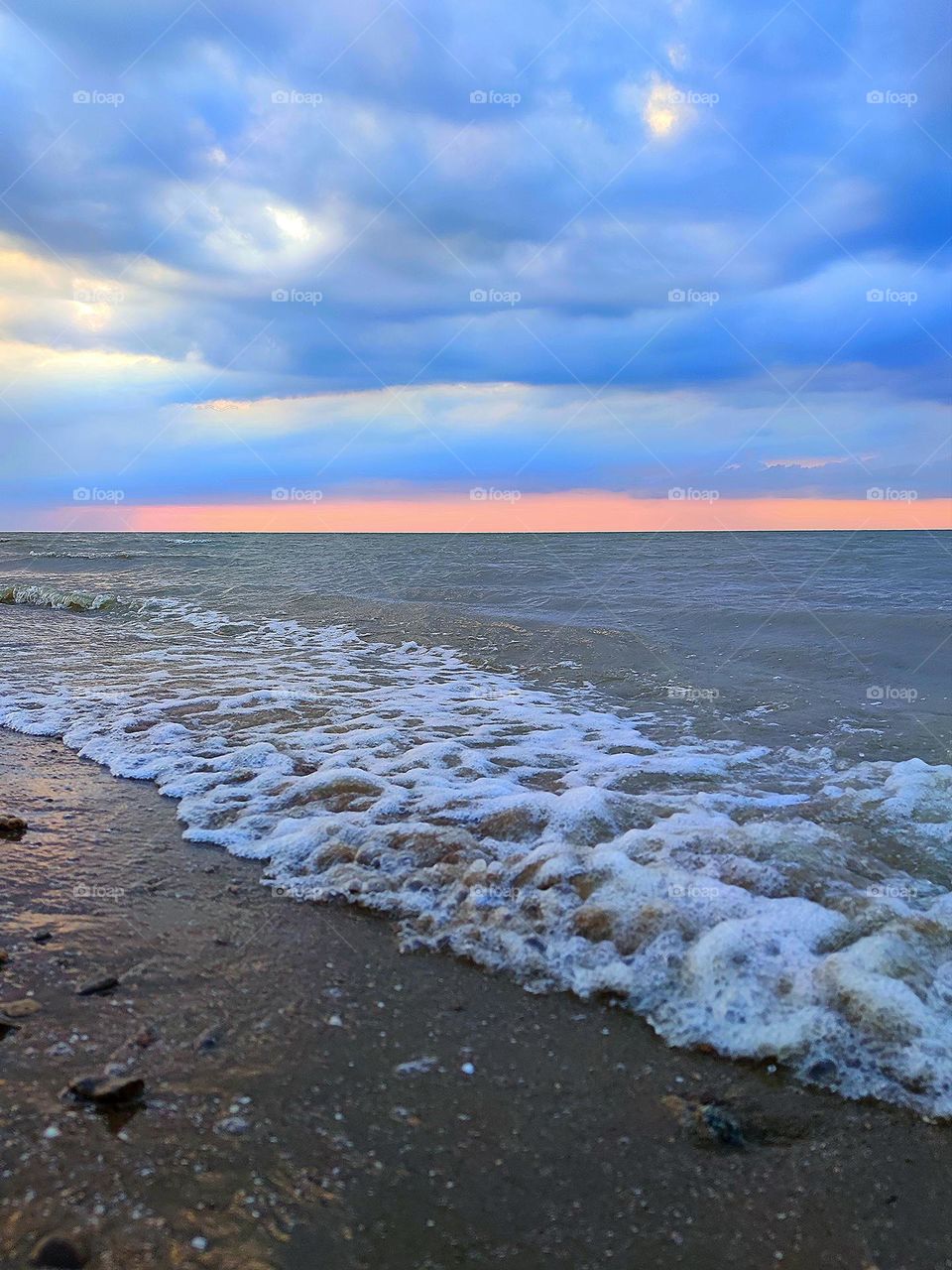 Summer sea.  Calm on the sea, small waves turn white near the shore.  The sunset colors the clouds in the sky blue.  The sky is tinted pink on the horizon