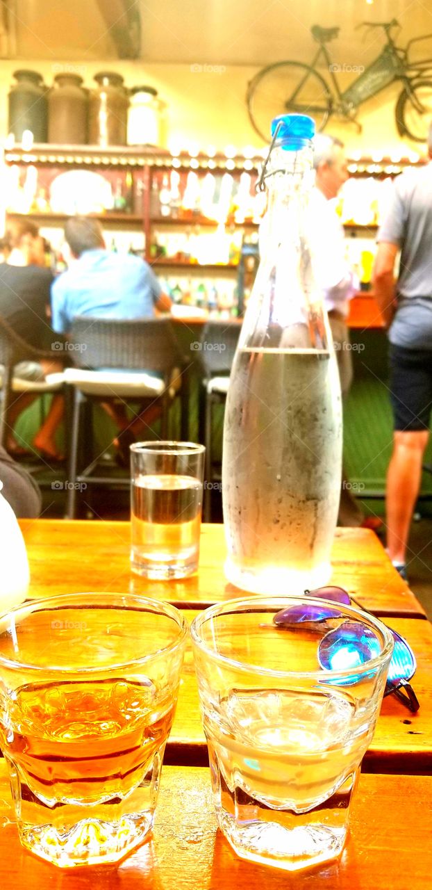 Perfect Summer set up at the neighborhood pub with neat whiskey & scotch cocktails on wood bar top with sunglasses to the side and quencher carafe of cold water.