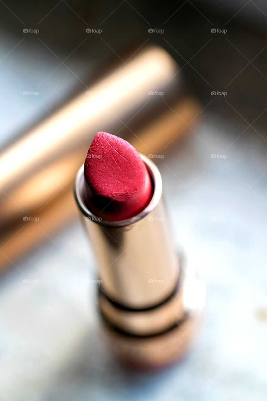 A portrait of a red lipstick in a gold tube container. there are some marks of use on it. the lit of the container is blurred in the background.