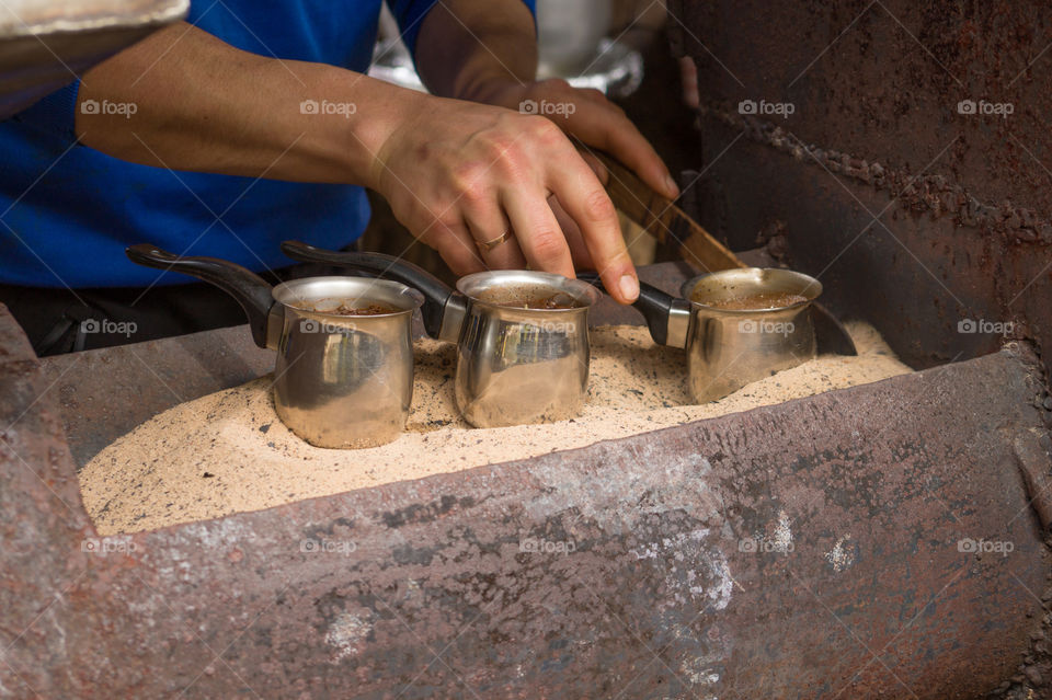 A man makes coffee on hot sand in a stainless turk