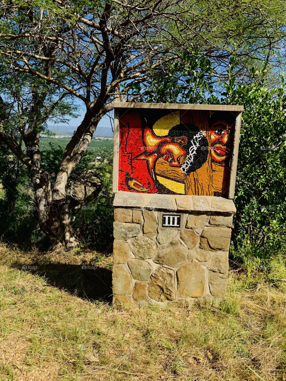 A rock crafted painting with bright colors. Roman numbered 3. On a designed rocky stand in the forest. 