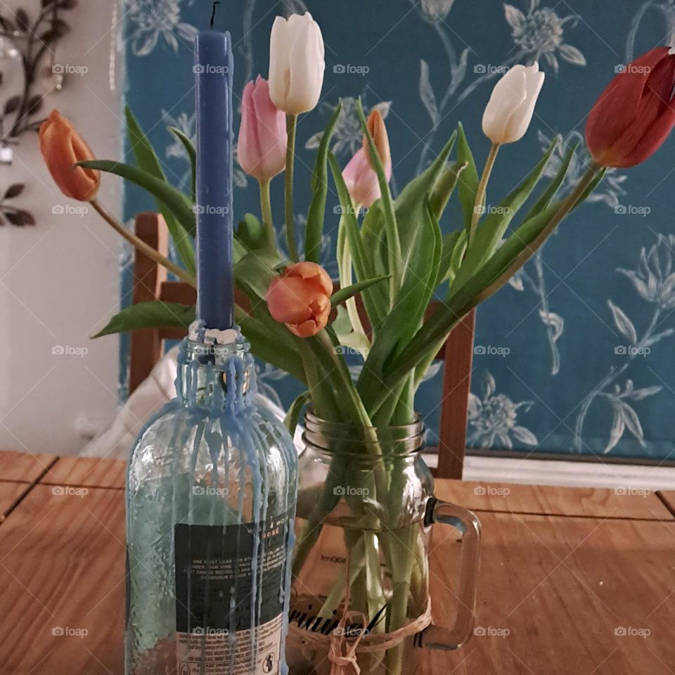 wooden table holding a bottle with a blue candle and a glass vase of bright tulips against a bluegreen windowblind