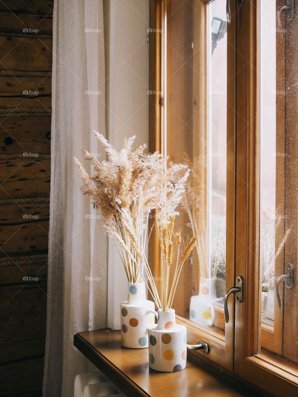 Branches of dried flowers stand in polka-dotted ceramic vases on a wooden window sill.